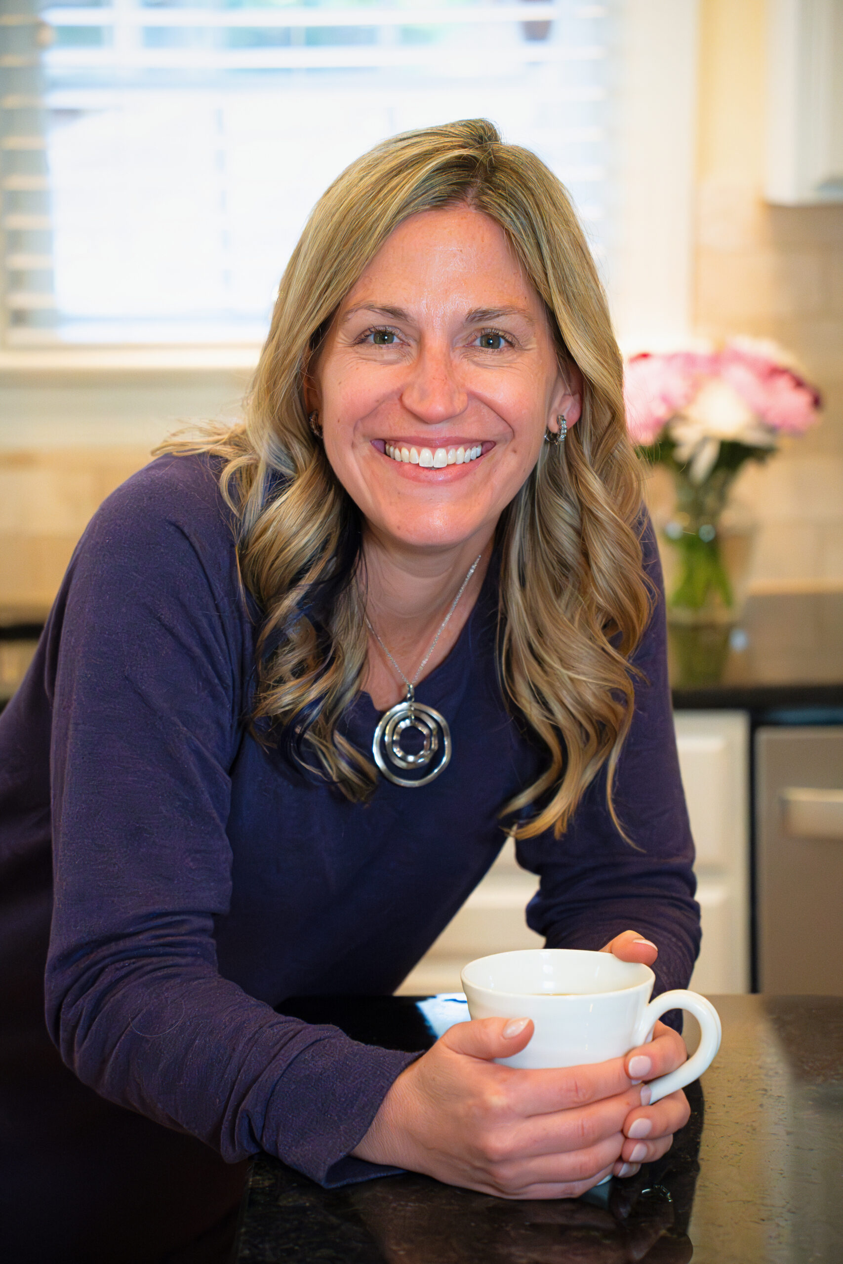 A woman in a purple sweater leaning over a counter and holding a white coffee mug.