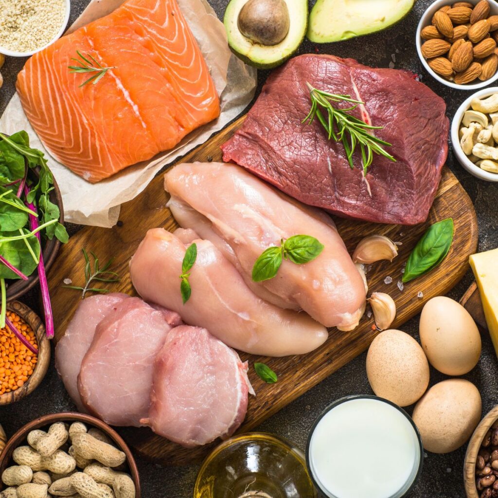 A collection of various protein sources grouped together on a cutting board and in bowls, including salmon, beef, chicken, pork chops, eggs, milk and nuts.